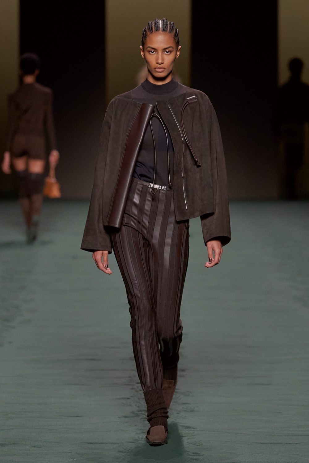 Runway model walks fashion show, wearing brown jacket, vertical-striped leather pants, black top. Another model visible in background.