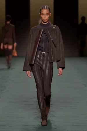 Runway model walks fashion show, wearing brown jacket, vertical-striped leather pants, black top. Another model visible in background.