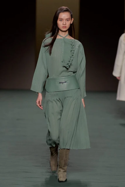 A runway model presents a monochrome sage green outfit featuring a pleated top, wide belt, and culottes during a fashion show.