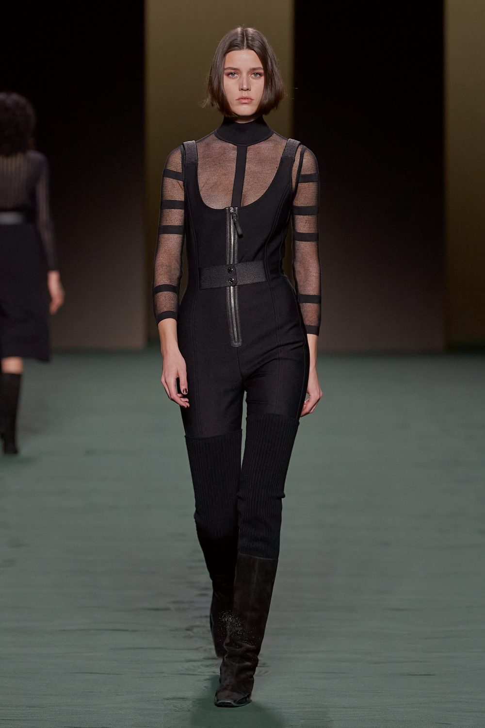 Runway fashion show features a model in a black jumpsuit with sheer sleeves and thigh-high boots walking on a green runway.