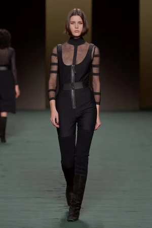 Runway fashion show features a model in a black jumpsuit with sheer sleeves and thigh-high boots walking on a green runway.