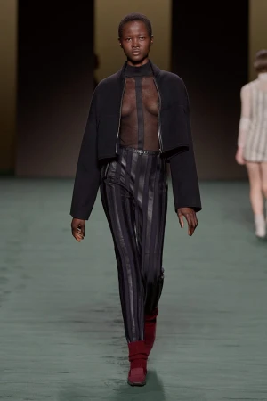 Runway fashion show featuring a model in a black jacket, sheer top, striped pants, and maroon boots, walking on a green runway.