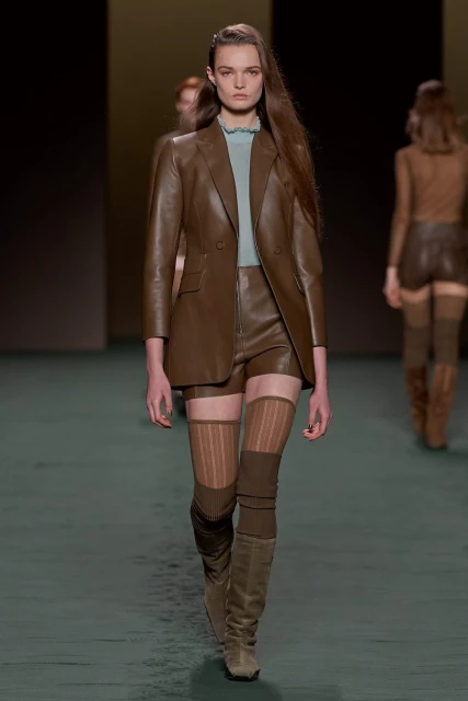 Runway fashion show featuring a female model in a brown leather blazer and shorts, paired with tan knit thigh-high socks and boots.