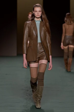 Runway fashion show featuring a female model in a brown leather blazer and shorts, paired with tan knit thigh-high socks and boots.