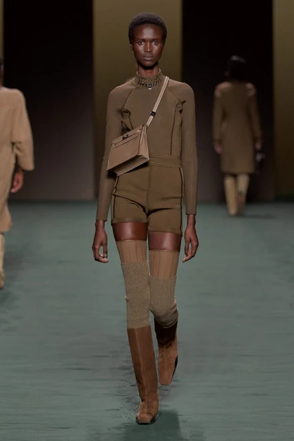 Runway fashion: A Black female model showcases a brown, long-sleeve top, shorts, boots, and crossbody bag on the catwalk.