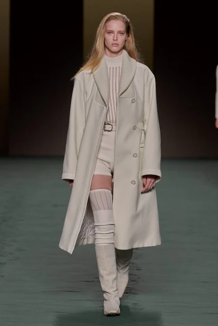 Runway fashion: Woman models a cream-colored coat, beige shorts, and knee-high boots in a studio setting with a muted background.