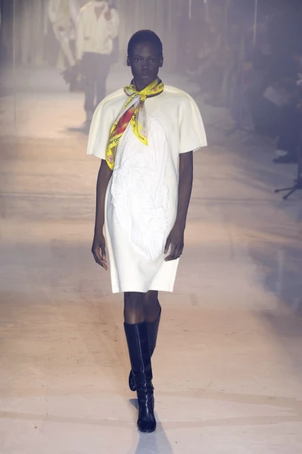 Runway shot of a Black female fashion model wearing a white dress, scarf, and black boots walking forward with lights and people in the background.