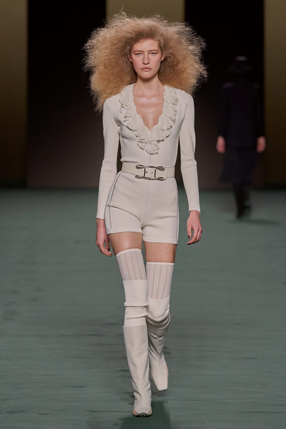 A fashion runway photograph featuring a model with large blonde afro wearing a cream-colored outfit and thigh-high boots in a neutral setting.