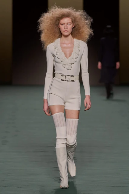 A fashion runway photograph featuring a model with large blonde afro wearing a cream-colored outfit and thigh-high boots in a neutral setting.