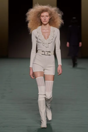A fashion runway photograph featuring a model with large blonde afro wearing a cream-colored outfit and thigh-high boots in a neutral setting.