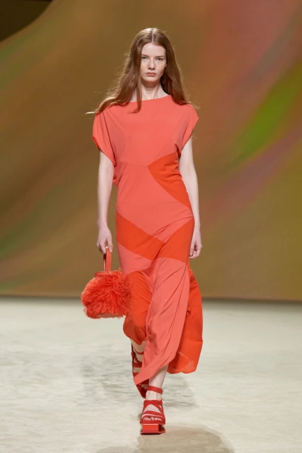 Runway fashion model in a coral dress and matching heels and a handbag walks on a neutral runway during a fashion show.
