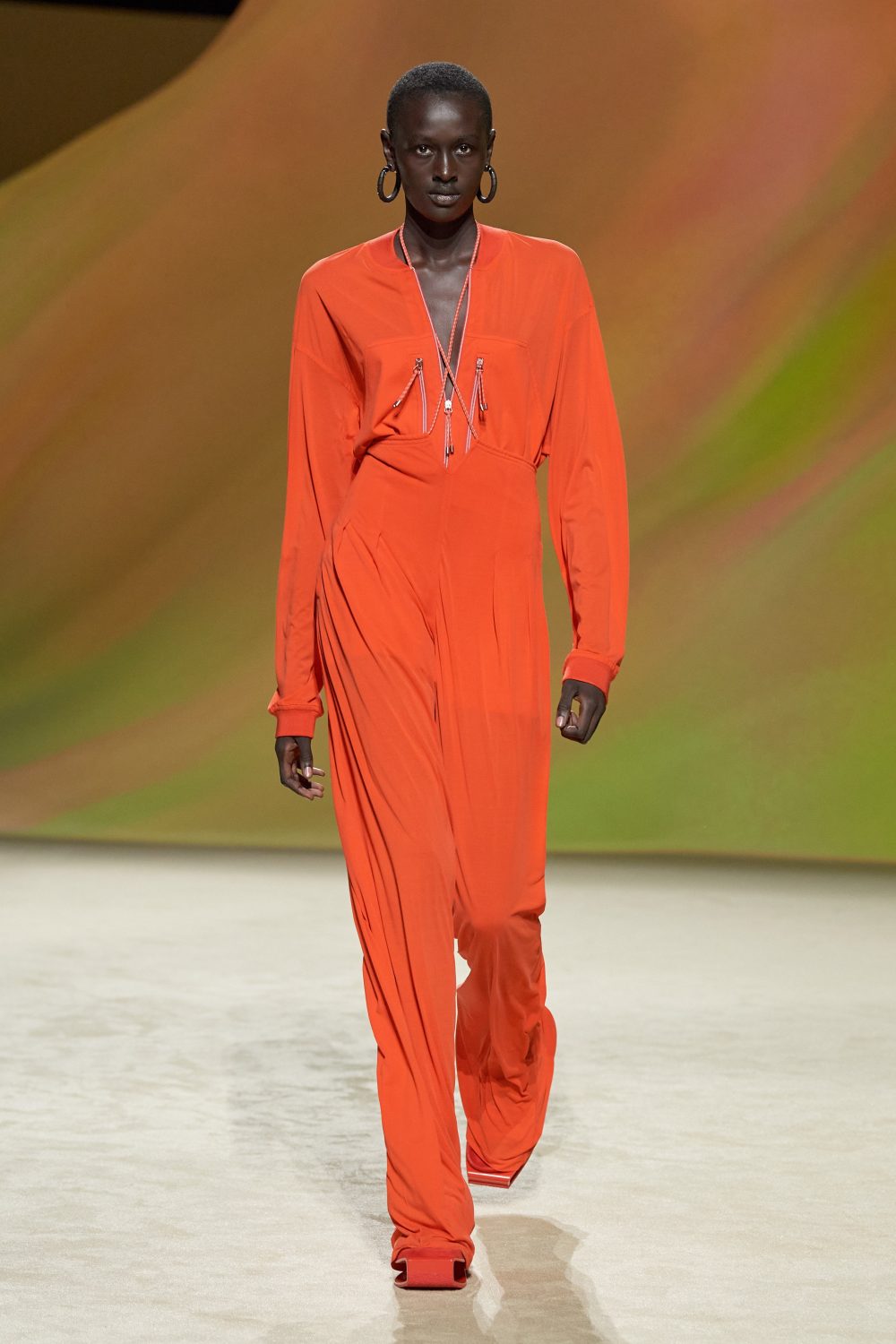 Runway fashion: a solo, slender Black woman strides confidently in a vibrant orange jumpsuit, hoop earrings, and a blurred backdrop.