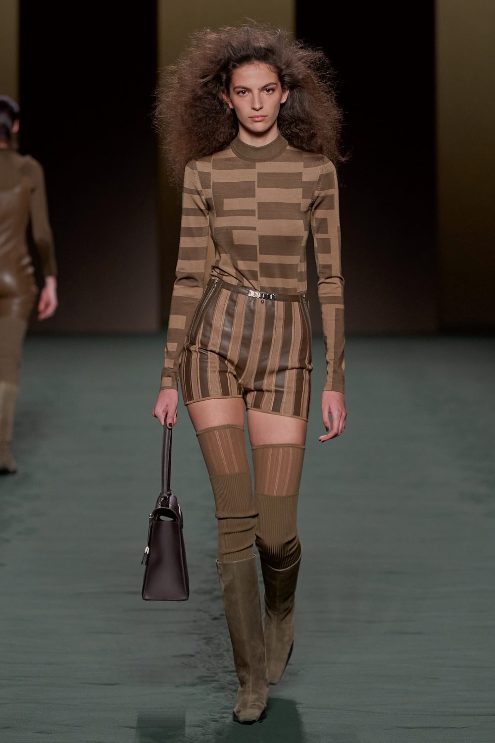 A fashion model walks the runway wearing a patterned top and shorts, brown boots, and carrying a dark brown handbag.