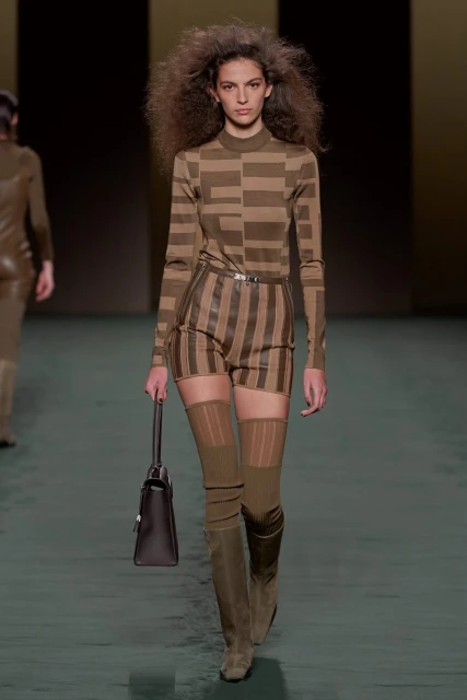 A fashion model walks the runway wearing a patterned top and shorts, brown boots, and carrying a dark brown handbag.