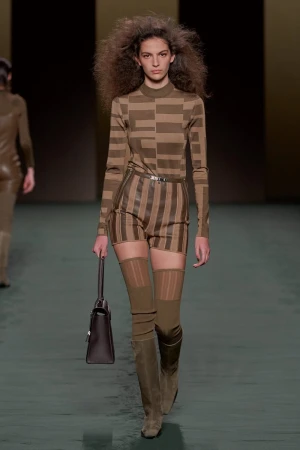 A fashion model walks the runway wearing a patterned top and shorts, brown boots, and carrying a dark brown handbag.