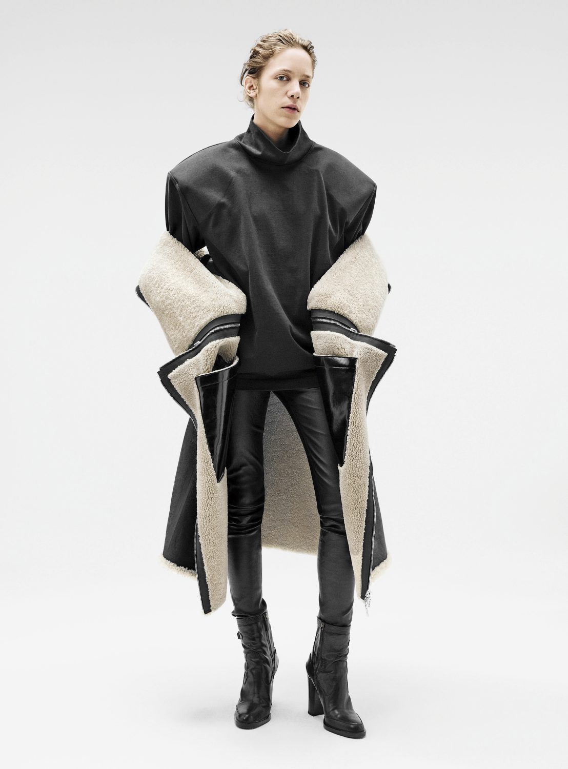 Model wearing a black oversized high-neck top, leather pants, and a shearling-lined coat draped around the shoulders against a plain background