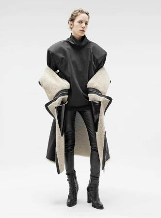 Model wearing a black oversized high-neck top, leather pants, and a shearling-lined coat draped around the shoulders against a plain background