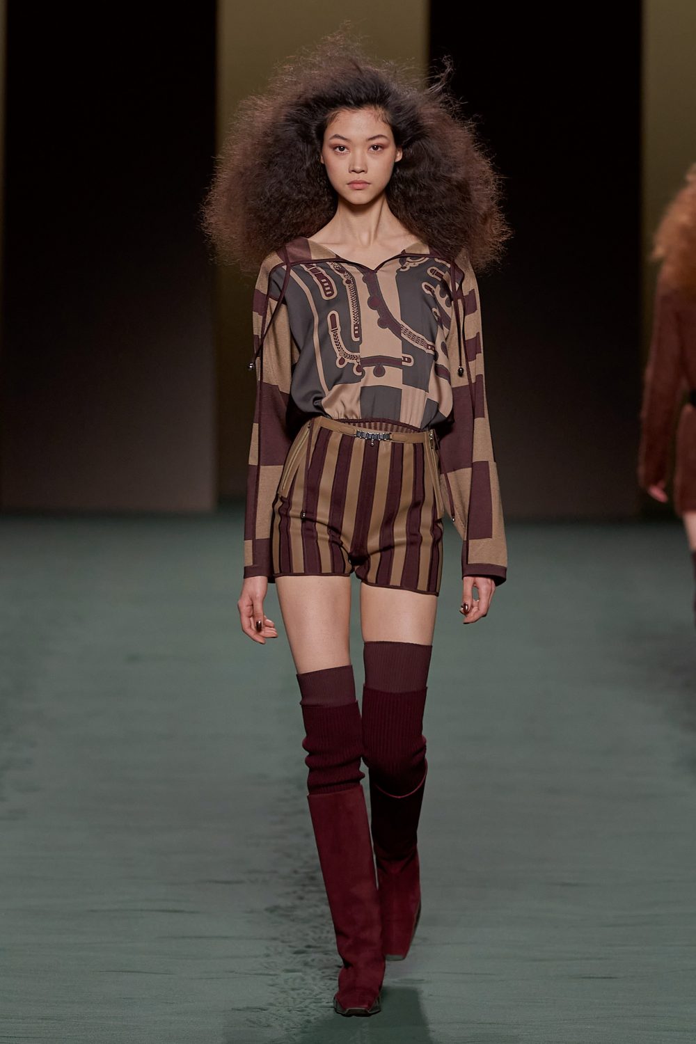 Runway fashion model showcases a brown and tan patterned long-sleeved top and striped shorts with thigh-high red suede boots on a runway.