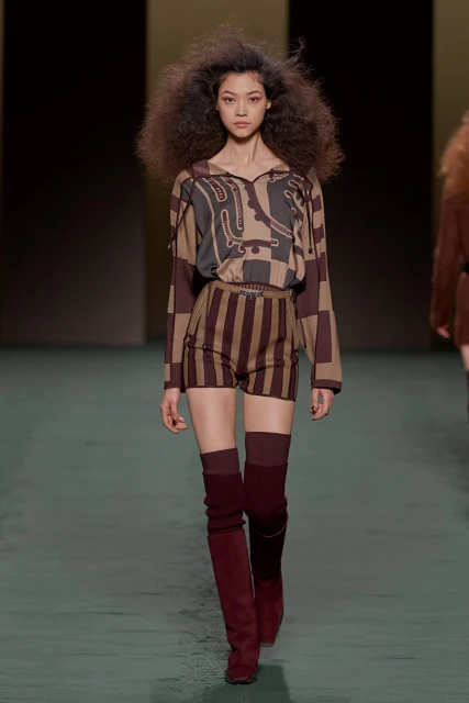 Runway fashion model showcases a brown and tan patterned long-sleeved top and striped shorts with thigh-high red suede boots on a runway.