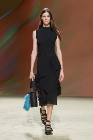 Runway model showcases a black sleeveless dress and black platform sandals, accessorized with a bucket bag with a blue feather charm.