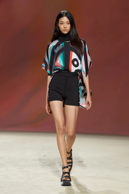 An Asian female fashion model walks the runway wearing a geometric print top, black shorts, and strappy sandals during a fashion show.