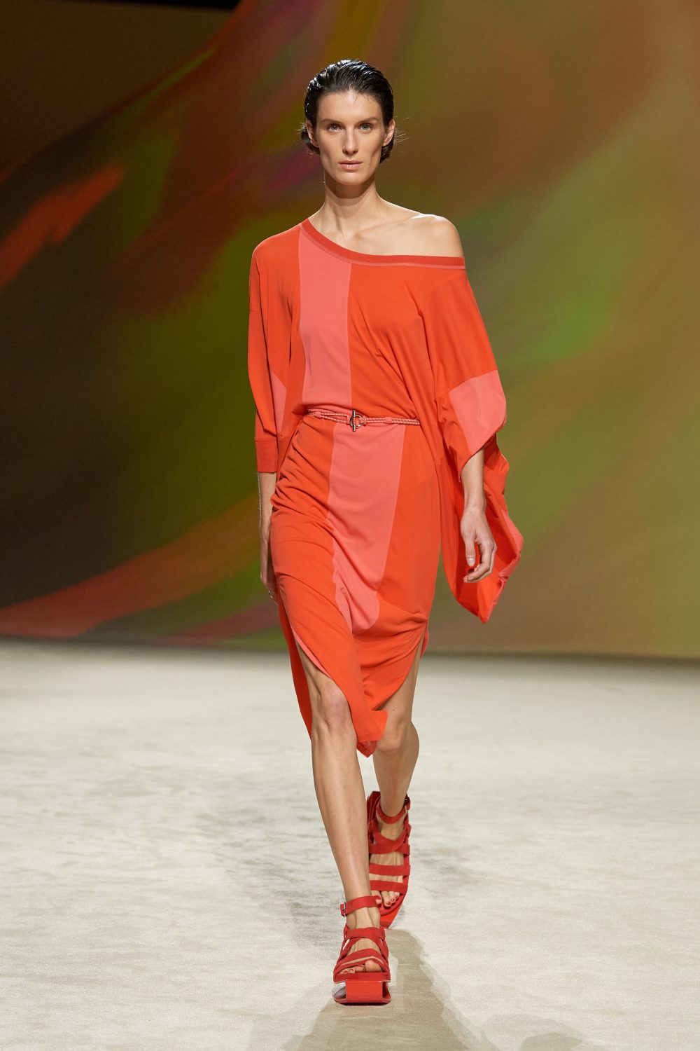 Runway fashion show featuring a model in an orange dress and red platform sandals, walking against a blended color background.