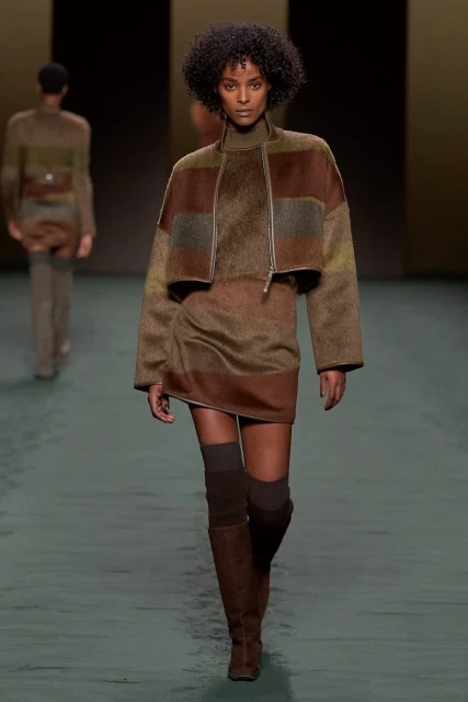 Female fashion model walks the runway in a brown and green striped jacket, matching mini dress, and over-the-knee boots, runway show.