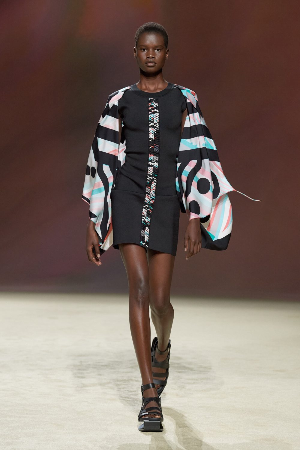 Runway shot of a woman in a black mini-dress with printed sleeves and gladiator platform sandals. The backdrop is a blended brown.