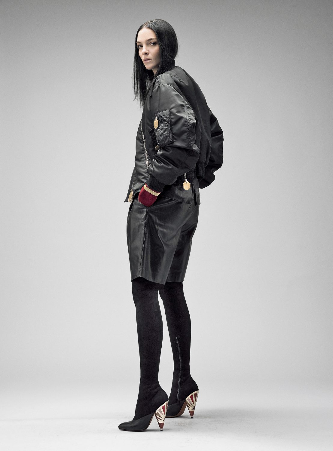 Fashion model poses in a black bomber jacket, leather shorts, and thigh-high boots against a plain background in a full-length studio shot.