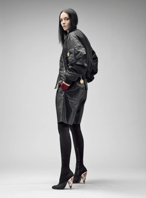 Fashion model poses in a black bomber jacket, leather shorts, and thigh-high boots against a plain background in a full-length studio shot.