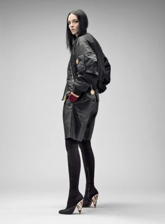Fashion model poses in a black bomber jacket, leather shorts, and thigh-high boots against a plain background in a full-length studio shot.