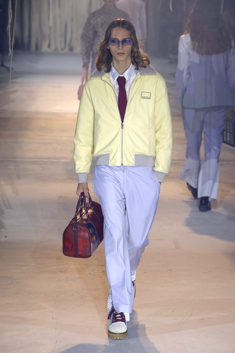 Fashion runway model wearing yellow jacket and light-blue pants, carrying a duffel bag, walking on the runway.
