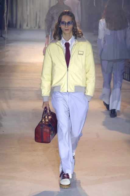 Fashion runway model wearing yellow jacket and light-blue pants, carrying a duffel bag, walking on the runway.