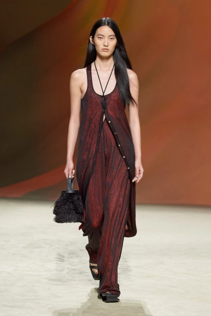 An Asian fashion model walks the runway in a sheer maroon dress and black sandals, carrying a black feathered handbag. Fashion show.