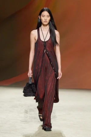 An Asian fashion model walks the runway in a sheer maroon dress and black sandals, carrying a black feathered handbag. Fashion show.