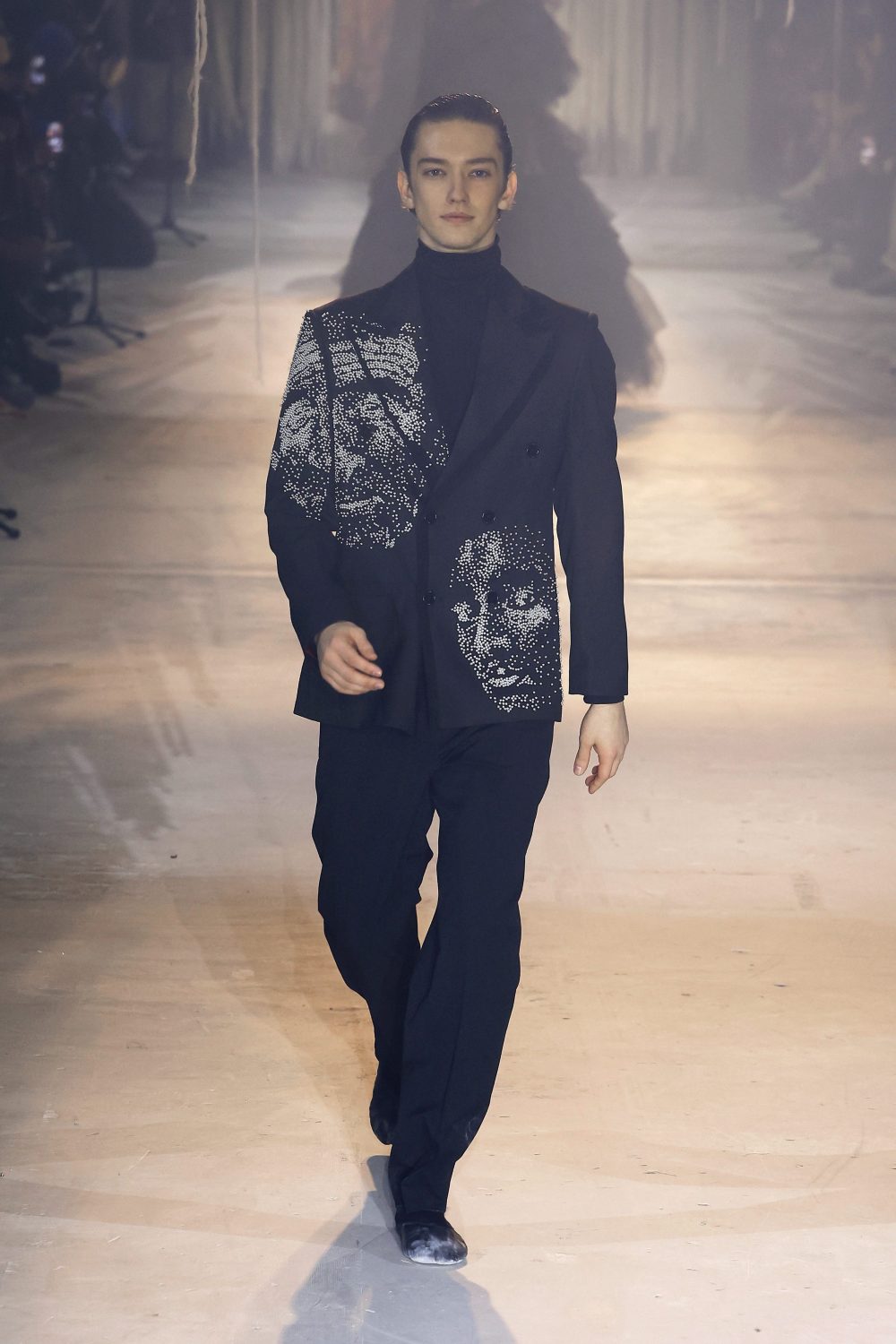 Runway model wearing a black suit with face graphics, black turtleneck, and dress shoes walking on a stage. Fashion show lighting.
