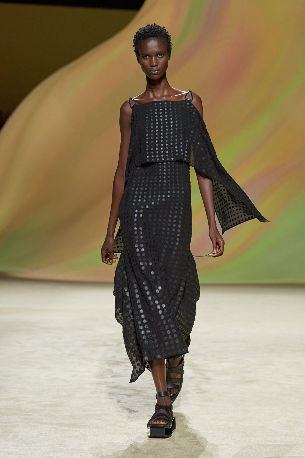Model on a runway wearing a black patterned dress with draped capelets and strappy sandals