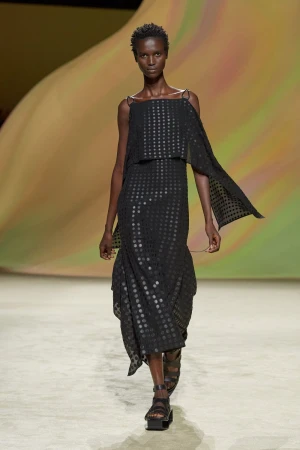 Model on a runway wearing a black patterned dress with draped capelets and strappy sandals