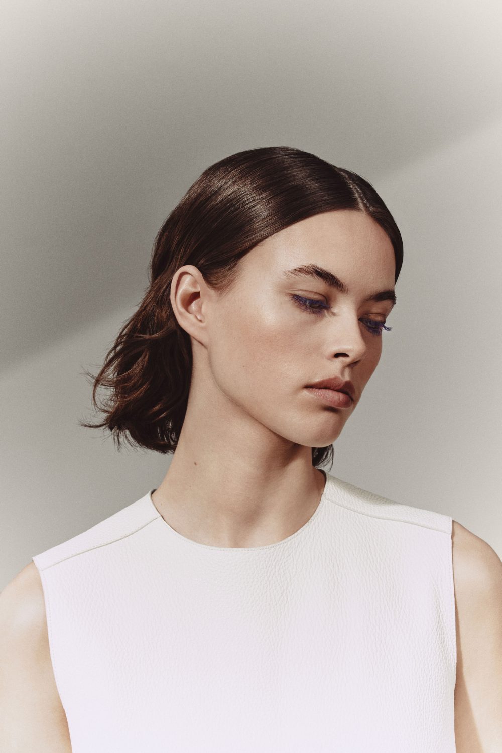 Close-up portrait of a young adult woman with dark hair, wearing a white sleeveless top against a neutral background in studio lighting.