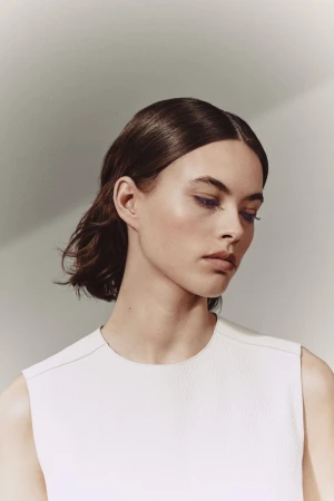 Close-up portrait of a young adult woman with dark hair, wearing a white sleeveless top against a neutral background in studio lighting.
