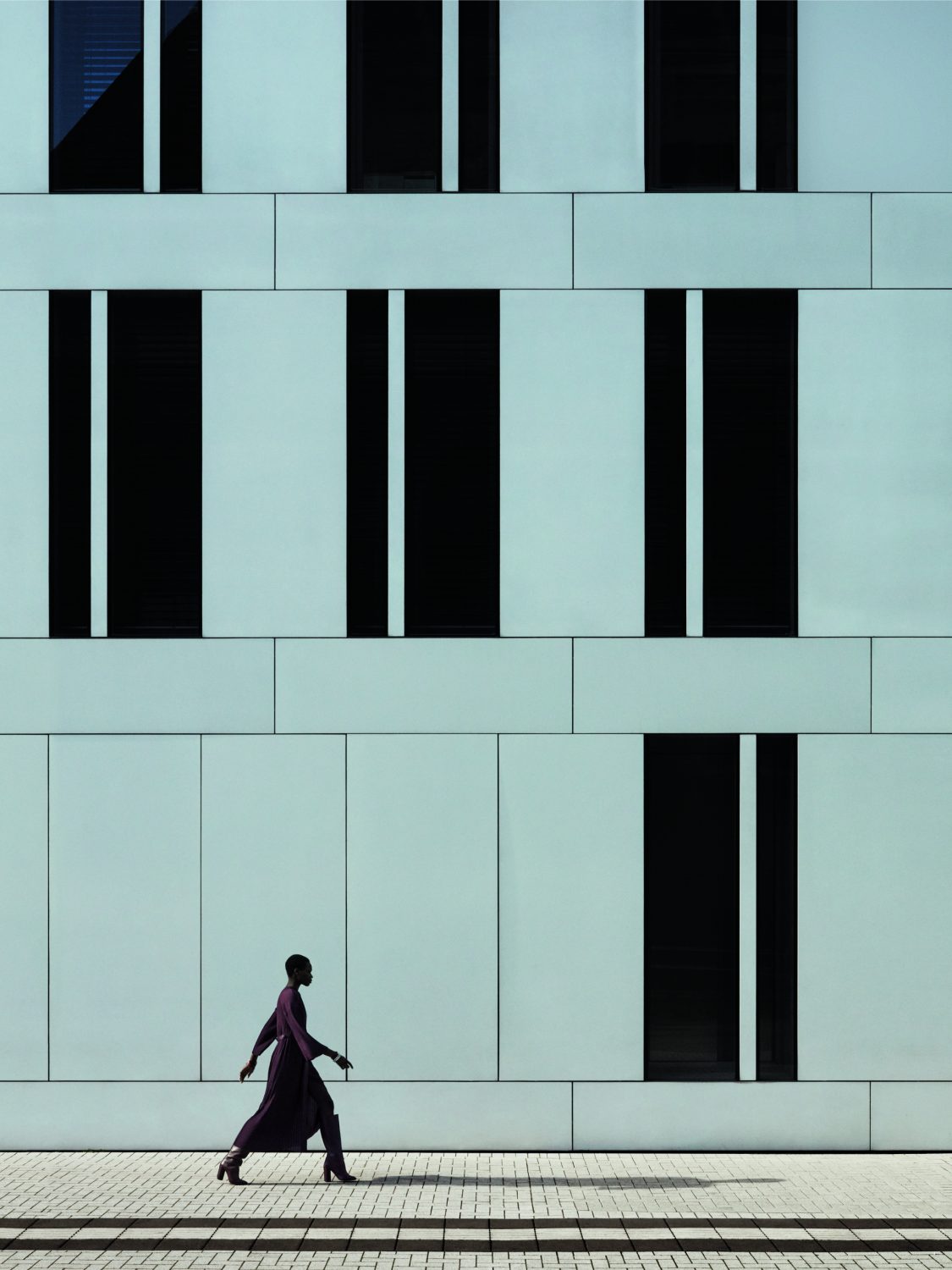 An adult female walking in a purple dress and high heels, posing outdoors in front of a blue building with black window accents.