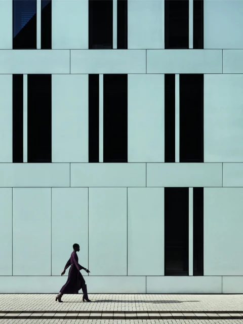 An adult female walking in a purple dress and high heels, posing outdoors in front of a blue building with black window accents.