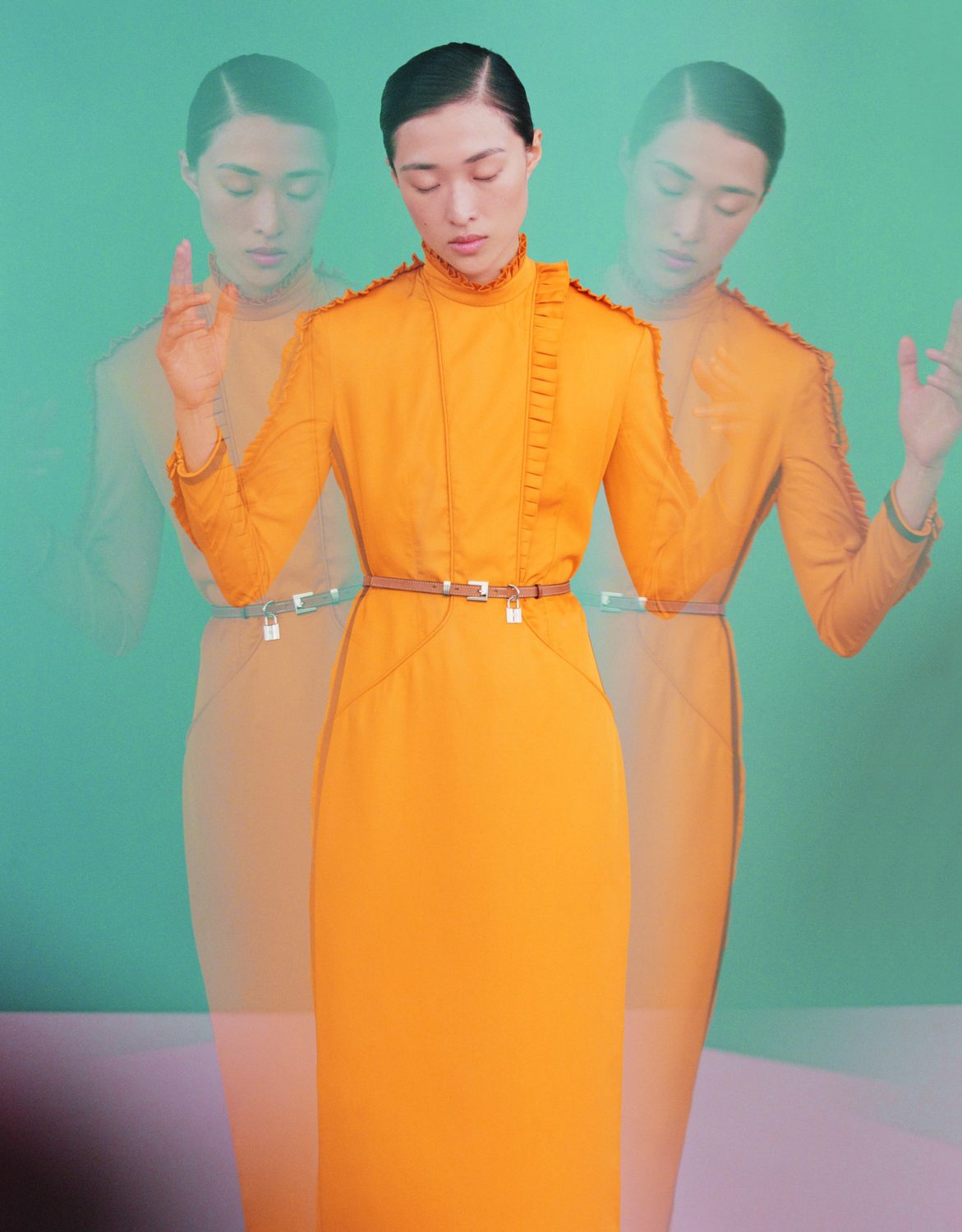 Woman wearing an orange dress posing in front of a mint green background, blurred duplicates of her in the background.