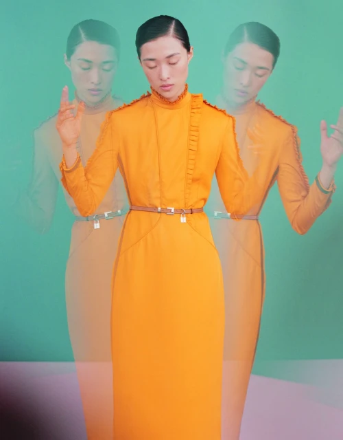 Woman wearing an orange dress posing in front of a mint green background, blurred duplicates of her in the background.