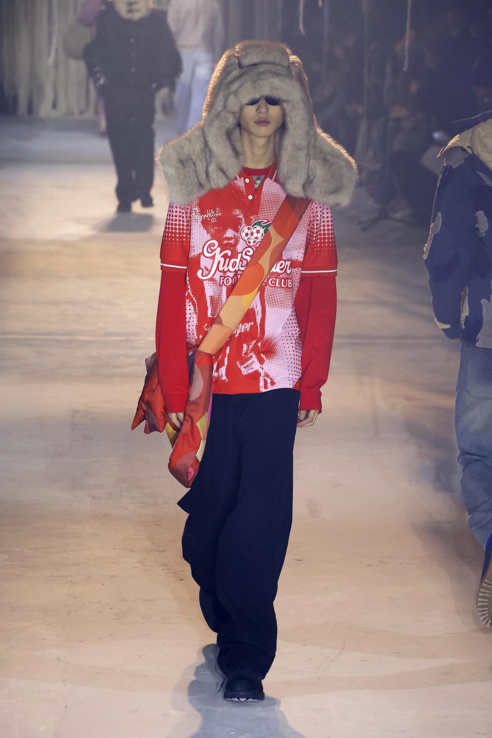Runway fashion show features a model in a red printed top and oversized fur hat, walking on a pale surface with audience members in the background.