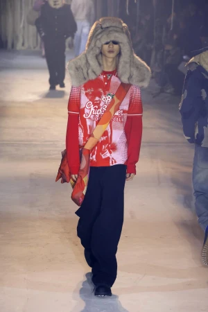 Runway fashion show features a model in a red printed top and oversized fur hat, walking on a pale surface with audience members in the background.