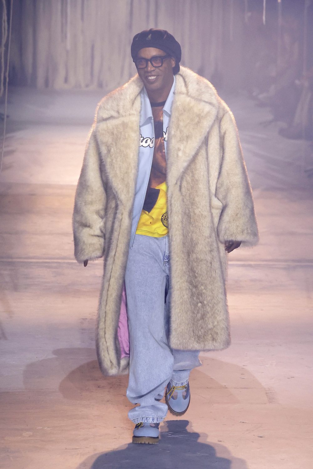 Man walks runway in oversized faux fur coat, light blue jeans, graphic shirt, and sneakers, wearing glasses and a black beret.