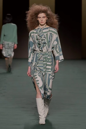 Model walking the runway wearing a patterned midi dress and tall white boots with voluminous hair.