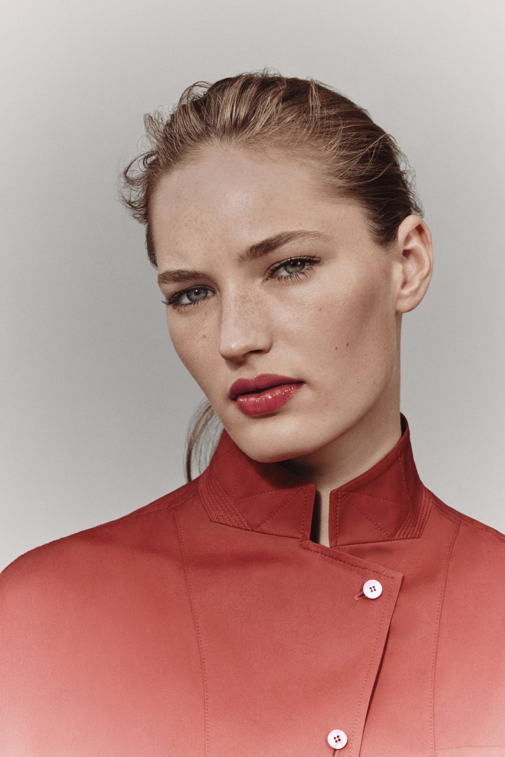 Close-up studio portrait of a blonde adult female model with red lipstick and a red shirt, set against a neutral grey background.