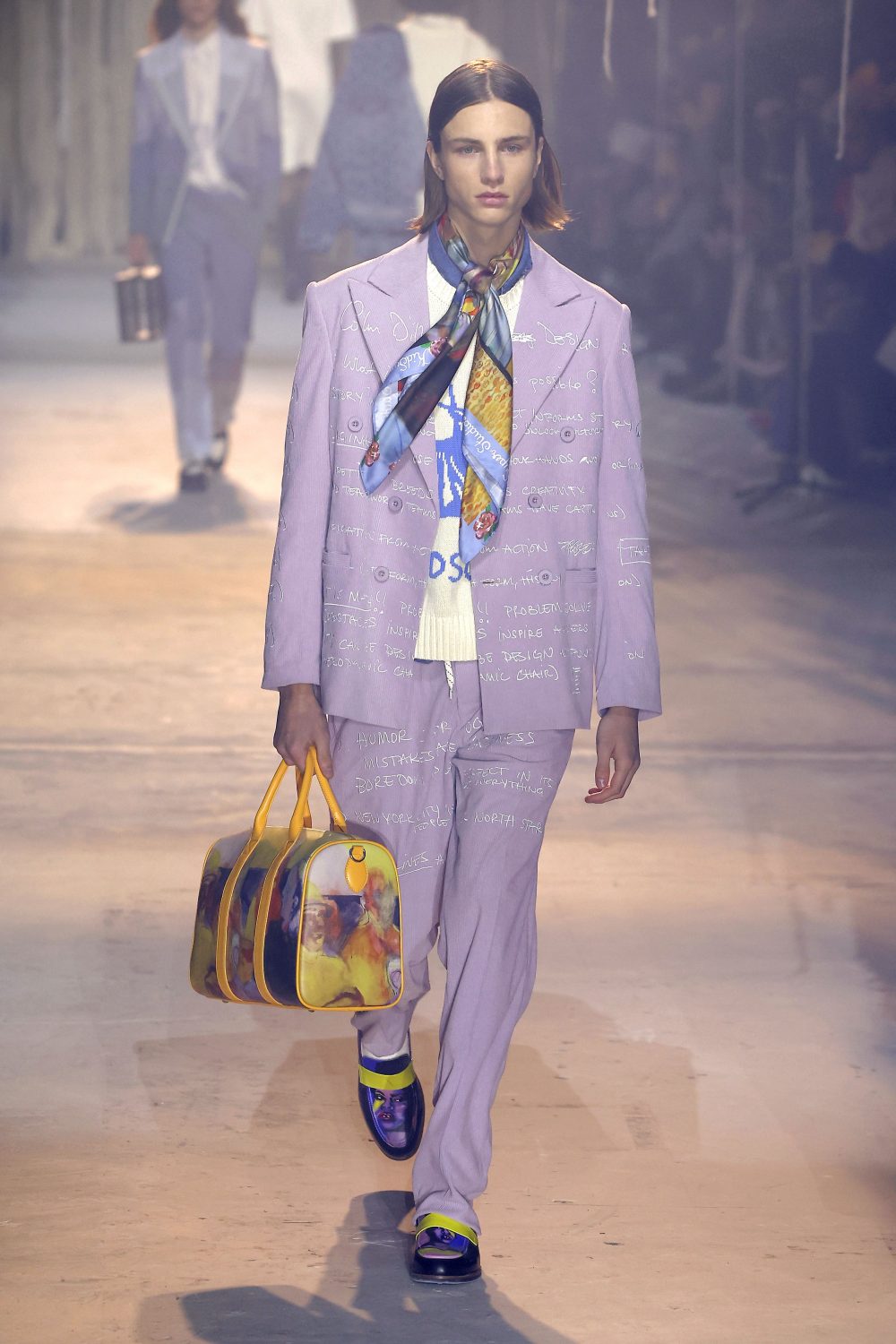Male fashion model wearing a lavender suit and carrying a duffel bag on a runway during a fashion show.